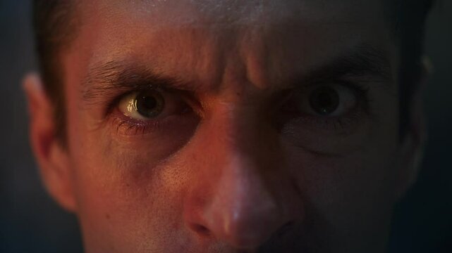 Close-up of angry man's eyes staring with anger at night illuminated by red light. Horror scene aggressive psychopath looking at camera. Macro shot angry male eyes close-up