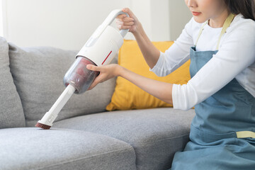 Cleaning people concept, happy smile asian young woman hand holding vacuuming dust and dirt on sofa, doing work chores cleanup furniture in living room at home. House clean with handheld hoover.