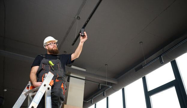 Man install a spot track LED light in the ceiling at the new office. Electrician staying on ladder. Industrial black loft design. Bearded man installing track light at the new apartment
