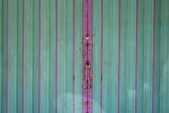A close-up view of a dilapidated, closed garage door in a green tint with purple iron bars dividing it, featuring three locks