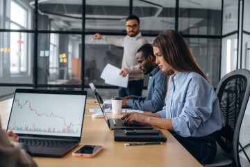Man is standing by the glass, showing information. Professional workers are in the modern office together