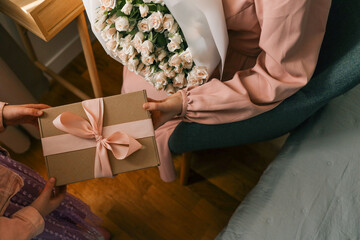 Close-up of a child giving a woman a gift box with a pink ribbon and a bouquet of delicate roses in...