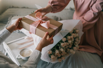 A wrapped gift with pink ribbon being handed over near a bouquet of roses and a cup of coffee on a...