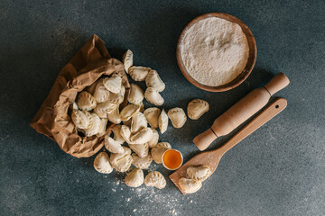 Hands holding a paper bag filled with homemade frozen dumplings on a kitchen counter, surrounded by flour, egg yolk, rolling pin, and wooden utensils