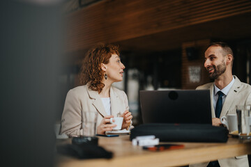 A man and woman discuss work over coffee in an outdoor setting, using a laptop for collaboration....