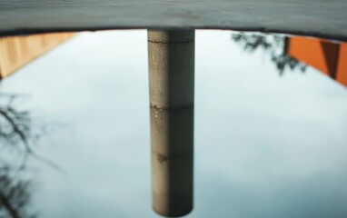 Fototapeta premium Concrete pillar reflected in a puddle, with muted colors and a cloudy sky in the background. The image is taken from a low angle, showing the reflection distorted by the water