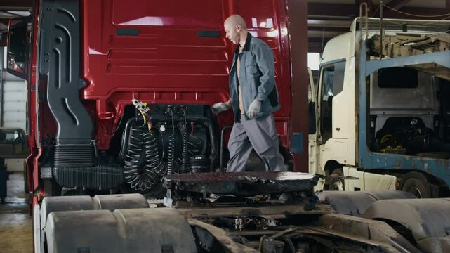 Full shot of middle-aged Caucasian male mechanic in workwear climbing red semi-truck for checkup, working in professional auto repair shop