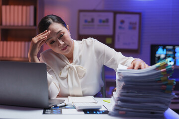 Stressed businesswoman staring at a towering stack of paperwork while working late at night in an...