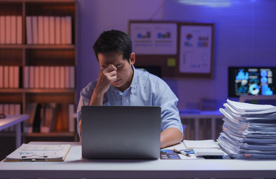 Stressed asian businessman is massaging his nose and having a headache from working late at night in office using laptop while sitting at desk with stacks of paperwork