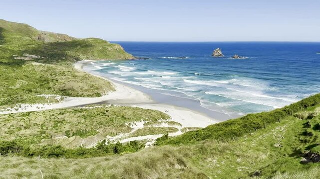 view of stunning idyllic sandfly bay. Powerful waves crashing on islands and white sand beach, hills covered with green bush. Otago Peninsula, New Zealand, South Island. Most beautiful beaches in  NZ