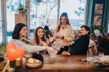 A cheerful group of young adults spending time together in a warm and inviting cafe, enjoying their interaction around a wooden table surrounded by cozy decor and natural light.