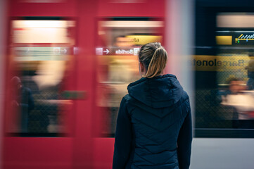A conceptual shot of a young blonde girl with a ponytail in a black jacket waiting at a metro station platform, as the train speeds by in Barcelona, capturing the urban vibe and motion.