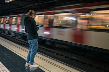A young woman in sportswear stands on a metro station platform in Barcelona, using her smartphone while waiting for the train to arrive.