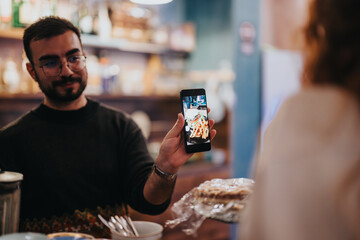 A man shares a picture of a meal on his phone with others in a cozy restaurant, highlighting friendship, food appreciation, and social moments in a vibrant atmosphere.