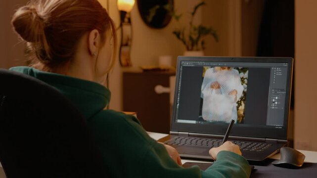 Woman is engaged in the creative process of editing images on her laptop using a graphics tablet. Her home studio is warm and inviting, showcasing her design skills.
