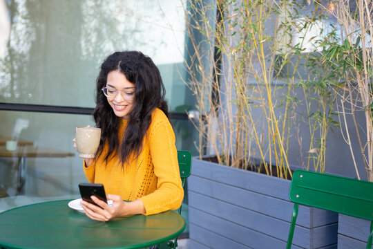 Latin woman using smartphone and drinking coffee at outdoor cafe