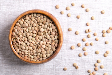 Large green lentils in a wooden bowl on linen fabric. Unshelled, dried plate lentils, a typically larger, flat variety of lentils, that hold their shape when cooked, used in soups, stews, or salads.