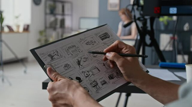 Slow motion shot of unrecognizable hand drawing with pencil on clipboard with gridded storyboard, while filming video on professional set
