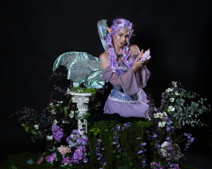 full length portrait of pretty female model wearing fantasy elf costume with fairy wings and purple dress. sitting pose, hands casting a magical spell, magical dark studio background