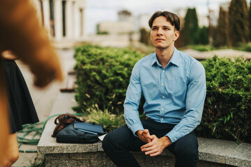 Fototapeta premium A young man in a blue shirt sits outdoors in an urban park, suggesting a casual business meeting. Lush greenery and city structures create a professional yet relaxed atmosphere.