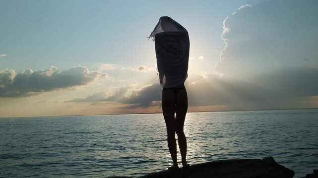 Grace silhouette of a beautiful bikini woman dancing by the sea at sunset. Young pareo girl standing on the beach with setting sun on the background.