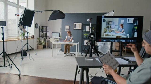 Woman in white shirt and gray vest using clapperboard, while director looking through camera as young woman acting in office scene amidst plants, books, and lighting equipment on brightly lit film set