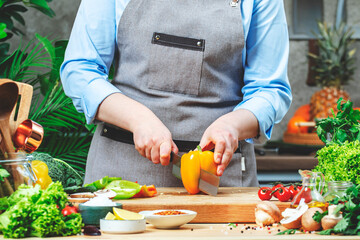 Chef blogger cuts orange paprika. Cozy kitchen with wooden table, kitchenware, vegetables, herbs and ingredients for cooking. Healthy vegan eating, culinary, recipes, food blogging