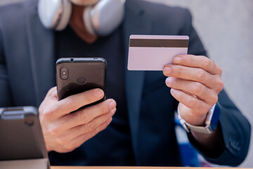 unrecognizable businessman in black suit using smartphone and holding credit card at outdoor cafe. Headphones around neck, tablet and coffee on table. Focused on making online payment or purchase. 