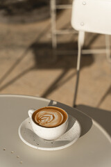 A latte with leaf-shaped art sits on a table bathed in warm sunlight.