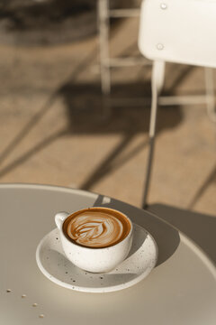 A latte with leaf-shaped art sits on a table bathed in warm sunlight.