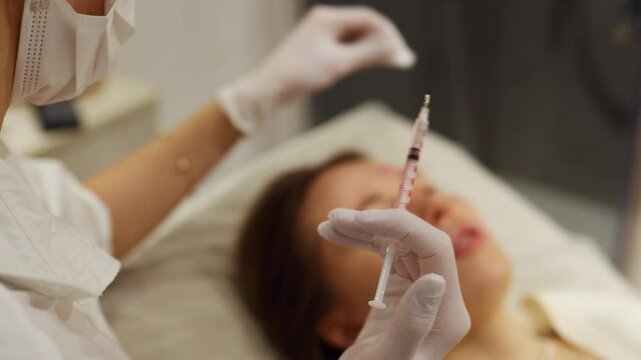 A woman relaxes as a professional administers a cosmetic treatment. Close-up of the doctor's hands injecting Botox into the girl's face and forehead with a syringe.