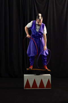 Circus artist balancing skillfully on a rola bola, holding two hoops in a dimly lit theater, showcasing precision and agility