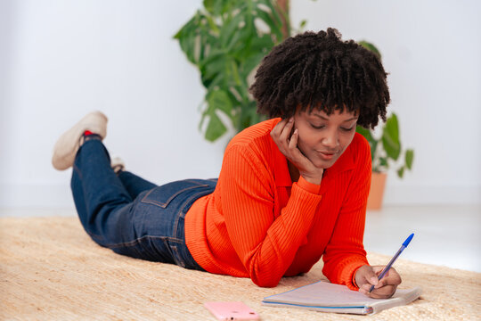 Relaxed african american young woman in orange sweater lying on carpeted floor and writing in a notebook. Peaceful home setting with green plant in background, casual and creative indoor atmosphere. 