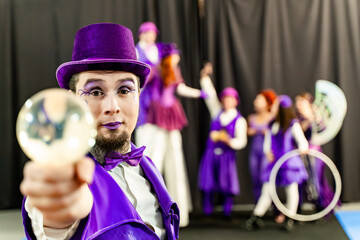 Magician wearing purple suit and top hat showing crystal ball with circus performers juggling and on stilts in background © JoseIMartin