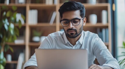 Young serious indian professional business man, focused ethnic male student wearing glasses working on laptop, remote studying using computer looking at screen watching seminar webinar at home office