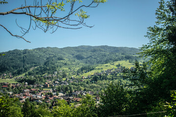 A scenic view of a mountain town surrounded by green hills and forests, with red-roofed houses in the valley. Trees and branches frame the composition, creating a calm, summery atmosphere.
