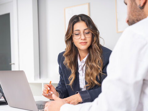 Two young busy caucasian professionals man and woman at meeting sitting desk at office using laptop checking documents reading financial report papers working together. Over shoulder view copy space.