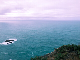 Ocean view from a cliffside with rocky shoreline and waves under a cloudy sky.
