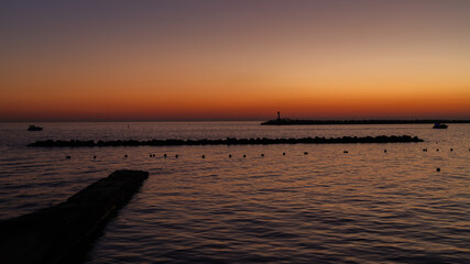 Tranquil sunset at sea with boats, gradual color transitions, breakwater, and lighthouse silhouettes, emanating peacefulness and tranquility.
