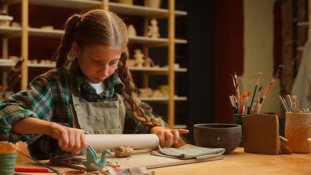 Young creative Caucasian little girl teen child junior kid schoolgirl student potter artist kneading clay material use rolling pin creating pottery artwork handmade ceramic art lesson inside workshop