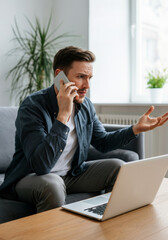 Frustrated young Caucasian man talking on phone with laptop in modern home office