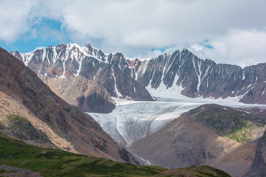 Scenic landscape with glacier tongue among rocky ridges and sharp snow-capped mountain range in sunlight under clouds in blue sky. Alpine valley with green hills and rocks against ice and sheer crags.