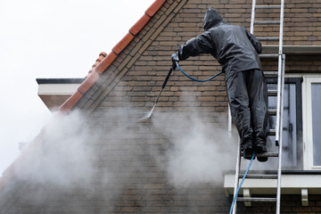 Man on ladder in waterproof suit cleans dirty brick facade of house with high pressure water jet. High pressure facade cleaning