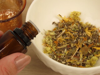 Fototapeta premium Woman hand holding a opened brown essential oil glass bottle over a white bowl with a mixture of dried herbs. Light wooden background. Self-care and wellness concept.