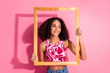 Smiling young woman holding a frame against a vibrant pink background, showcasing curly hair,...
