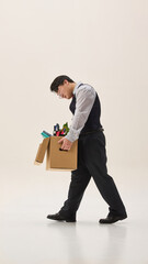 Young Asian man, dressed formal attire, leaving his job with heavy cardboard box, walking with sad expression against white background. Concept of human emotions, business, challenges. Ad