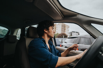 A man sits inside a parked car in an urban parking lot, holding car keys. He appears thoughtful, gazing out the window at surrounding buildings and vehicles. © qunica.com