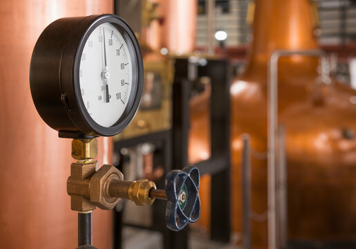 A copper still in a distillery, a large copper chamber and pipework and a large gauge and tap.
