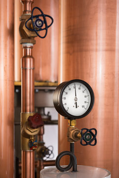 A copper still in a distillery, a large copper chamber and pipework and a large gauge and tap.