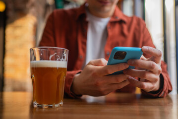 Hands holding smartphone with glass of beer on wooden table in pub, man browsing social media or texting messages while enjoying alcoholic beverage